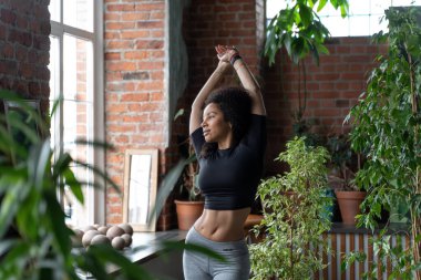 Slim Afro-American woman stretching at home or fitness studio in the morning. Studio decorated with plenty house plants, big windows and brick wall. Concept of happy and conscious healthy lifestyle.