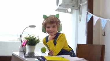 Cute smart caucasian primary School Child with headphones typing on the keyboard laptop. Online tutor teaching digital class, web lesson on computer at home. Virtual education. Schoolboy girl studying