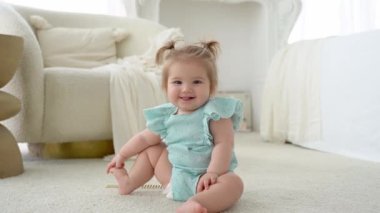 Little cute baby girl is sitting in a bright cozy bedroom floor smiling, looking at camera, showing tongue. Child makes funny faces. Babyhood, childhood, toddler lifestyle. Positive emotion. Carefree