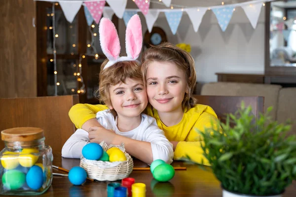 Easter Family traditions. Two caucasian happy children with bunny ears dye and decorate eggs with paints for Easter holidays while sitting together at home table. Kids enderly embracing and smiling in cozy light kitchen