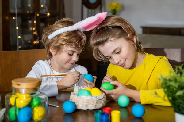 Easter Family traditions. Two caucasian happy children with bunny ears dye and decorate eggs with paints for holidays while sitting together at home table. Kids having fun together