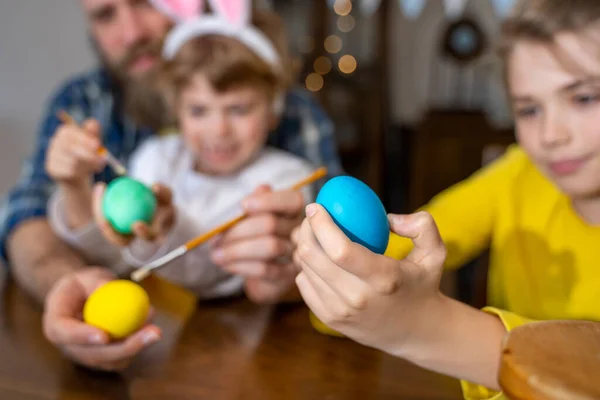 Easter Family traditions. Father and two caucasian happy children with bunny ears dye and decorate eggs with paints for holidays having fun together at home. Negative space