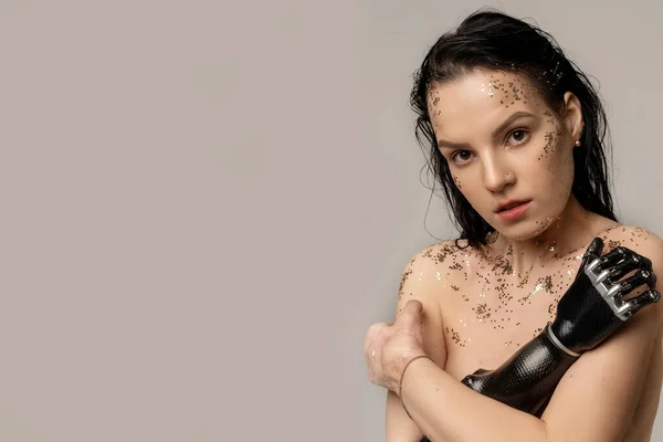 Slim Disabled Woman with Prosthetic Arm, Artificial Hand With Glitter Fresh Nude Makeup Looking At Camera Over Beige Background. Women Beauty Diversity. Positive. Copy Space