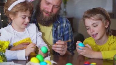 Easter Family traditions. Father and two caucasian happy children with bunny ears dye and decorate eggs with paints for holidays while sitting together at home table. Kids embrace and smile in cozy.