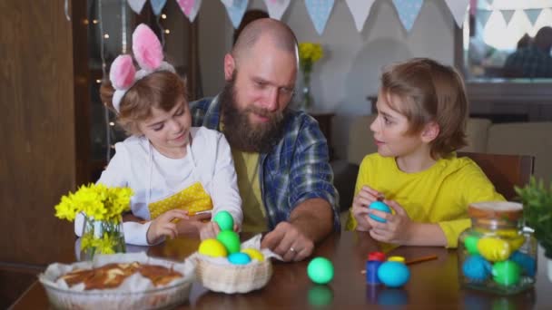 イースターファミリーの伝統 ウサギの耳を持つ父と2人の白人の幸せな子供は 家のテーブルに座っている間 休日のために塗料で卵を染め 子供たちは居心地の良いで受け入れると笑顔 — ストック動画