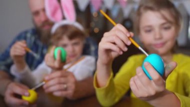 Easter Family traditions. Dad and two caucasian happy children with bunny ears dye and decorate eggs with paints for holidays while sitting together at home Easter Sunday table. Christian religious