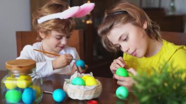 Easter Family traditions. Two caucasian happy siblings kids with bunny ears paint and decorate eggs with paints for holidays while sitting together at home table. Kids having fun together