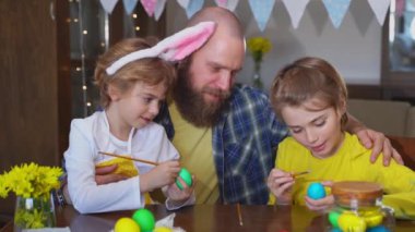 Easter Family traditions. Father and two caucasian happy children with bunny ears dye and decorate eggs with paints for holidays while sitting together at home table. Kids embrace and smile in cozy.