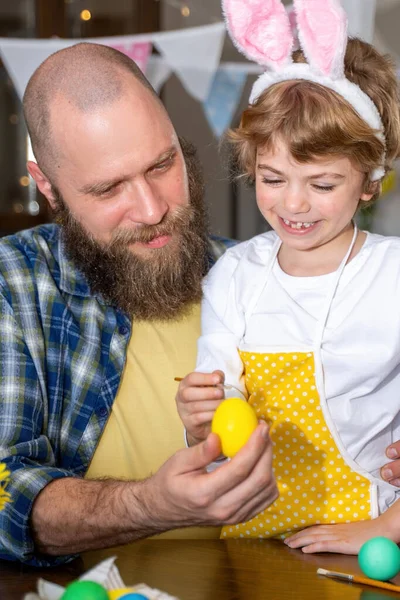 Easter Family traditions. Father and caucasian happy child with bunny ears dye and decorate eggs with paints for holidays while sitting together at home table. Vertical