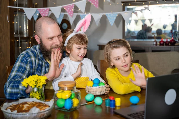 Easter Family traditions. Father and two caucasian happy children with bunny ears dye and decorate eggs with paints for holidays while sitting together at home table. Video call laptop with relatives