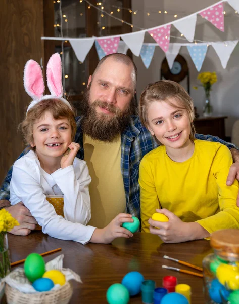 Easter Family traditions. Dad and two caucasian happy children with bunny ears play and decorate eggs with paints for holidays while sitting together at home table having fun together