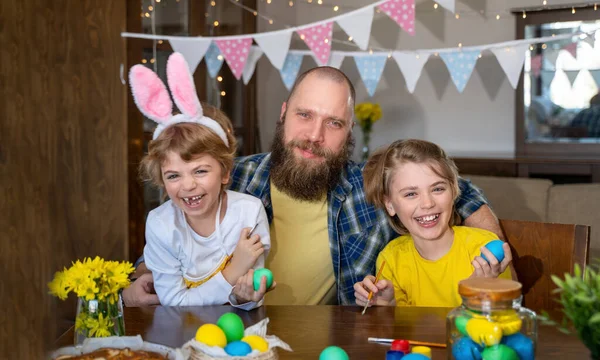 Easter Family traditions. Father and two caucasian happy smiling children with bunny ears dye and decorate eggs with paints for holidays while sitting together at home table. Dad embrace and smile in