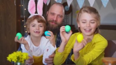 Easter Family traditions. Father and two caucasian happy children with bunny ears playing with Easter decorated eggs while sitting together at home holiday table.