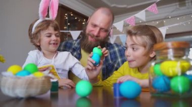 Easter Family traditions. Father and two caucasian happy children with bunny ears playing with Easter eggs with paints for holidays while sitting together at home table. Kids embrace and smile in cozy