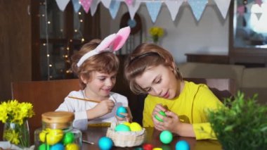 Easter Family traditions. Two caucasian happy school children kids with bunny ears paint and decorate eggs with paints for holidays while sitting together at home table. Kids having fun together
