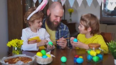 Easter Family traditions. Father and two caucasian happy children with bunny ears dye and decorate eggs with paints for holidays while sitting together at home table. Kids embrace and smile in cozy.