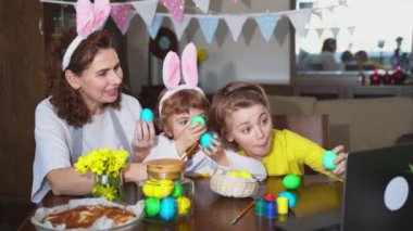 Easter Family traditions. Mom and two caucasian happy children with bunny ears dye and decorate eggs with paints for holidays while sitting together at home Easter Sunday table using laptop remote