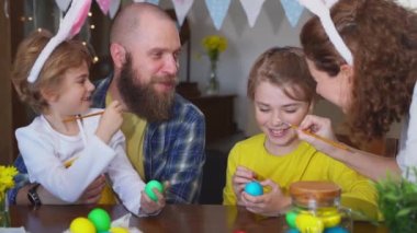 Easter Family traditions. Father and two caucasian happy children with bunny ears dye and decorate eggs with paints for holidays while sitting together at home table. Kids embrace and smile in cozy.