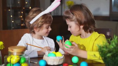 Easter Family traditions. Two caucasian happy siblings kids with bunny ears paint and decorate eggs with paints for holidays while sitting together at home table. Kids having fun together