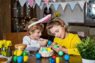 Easter Family traditions. Two caucasian happy children with bunny ears dye and decorate eggs with paints for holidays while sitting together at home table. Kids having fun together