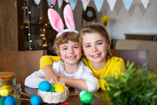 Easter Family traditions. Two caucasian happy children with bunny ears dye and decorate eggs with paints for holidays while sitting together at home table. Kids embrace having fun together