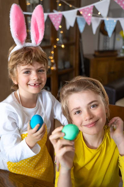 Easter Family traditions. Two caucasian happy children with bunny ears dye and decorate eggs with paints for holidays playing together. Kids having fun. Vertical