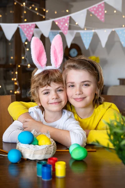 Easter Family traditions. Two caucasian happy children with bunny ears dye and decorate eggs with paints for holidays while sitting together at home table. Kids embrace and smile in cozy. Vertical