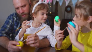 Easter Family traditions. Dad and two caucasian happy children with bunny ears dye and decorate eggs with paints for holidays while sitting together at home Easter Sunday table. Christian religious