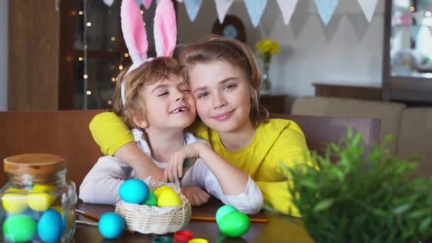 イースター日曜日家族の休日の伝統 2人の白人幸せな兄弟ウサギの耳を持つ子供たちは 家のテーブルに座っている間にカメラで飾られた卵を見て笑ってポーズをとっています 楽しい子供たち — ストック動画