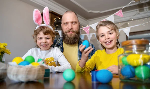 Paskalya Ailesi gelenekleri. Baba ve tavşan kulaklı iki Kafkasyalı mutlu çocuk tatil için yumurtaları boyayarak ev masasında birlikte oturuyorlar. Kameraya bakıyorum.