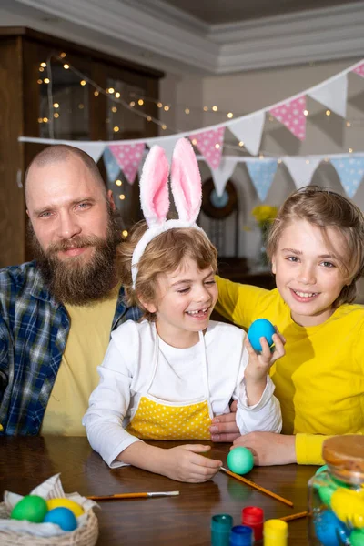 Easter Family traditions. Father and two caucasian happy children with bunny ears dye and decorate eggs with paints for holidays while sitting together at home table. Vertical
