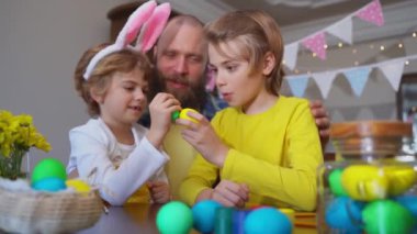 Easter Family traditions. Father and two caucasian happy children with bunny ears playing with Easter eggs with paints for holidays while sitting together at home table. Kids embrace and smile in cozy