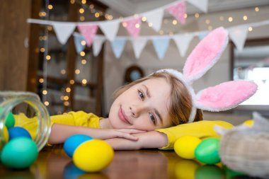 Easter Family traditions. Close-up caucasian child bunny ears playing with decorated multi-colored eggs. Kid having fun looking at camera. Negative copy space