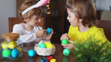 Easter Family traditions. Two caucasian happy siblings kids with bunny ears paint and decorate eggs with paints for holidays while sitting together at home table. Kids having fun together
