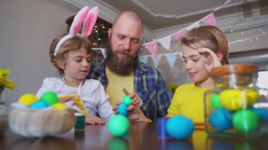 Easter Family traditions. Father and two caucasian happy children with bunny ears playing with Easter eggs with paints for holidays while sitting together at home table. Kids embrace and smile in cozy