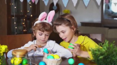 Easter Family traditions. Two caucasian happy children with bunny ears dye and decorate eggs with paints for holidays while sitting together at home table. Kids having fun together