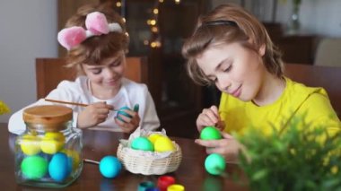 Easter Family traditions. Two caucasian happy school children kids with bunny ears paint and decorate eggs with paints for holidays while sitting together at home table. Kids having fun together