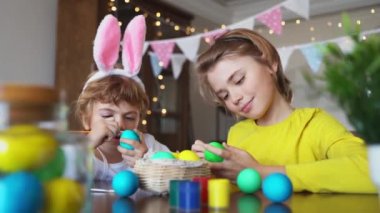 Easter Family traditions. Two caucasian happy siblings kids with bunny ears paint and decorate eggs with paints for holidays while sitting together at home table. Kids having fun together