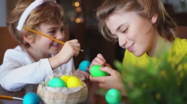 Easter Family traditions. Two caucasian happy siblings kids with bunny ears paint and decorate eggs with paints for holidays while sitting together at home table. Kids having fun together