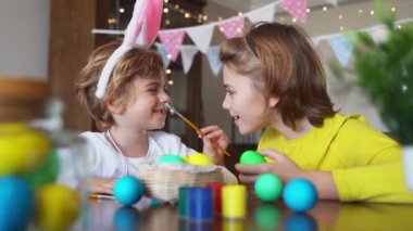 Easter Family traditions. Two caucasian happy siblings kids with bunny ears paint and decorate eggs with paints for holidays while sitting together at home table. Kids having fun together. Negative