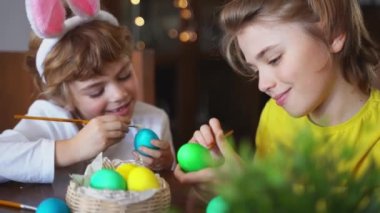 Easter Family traditions. Two caucasian happy siblings kids with bunny ears paint and decorate eggs with paints for holidays while sitting together at home table. Kids having fun together