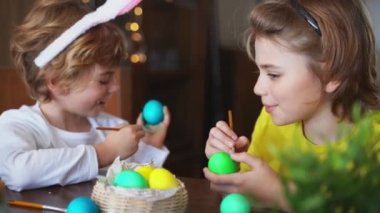 Easter Family traditions. Two caucasian happy school children kids with bunny ears paint and decorate eggs with paints for holidays while sitting together at home table. Kids having fun together