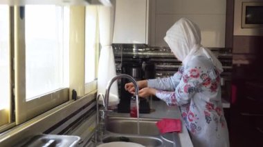 Orta yaşlı Müslüman kadın başörtüsü takarak ev işi yapıyor mutfak lavabosu temizliyor. İslami kadın ev bulaşıklarını yıkıyor. Ev hanımı günlük ev hayatı rutini.