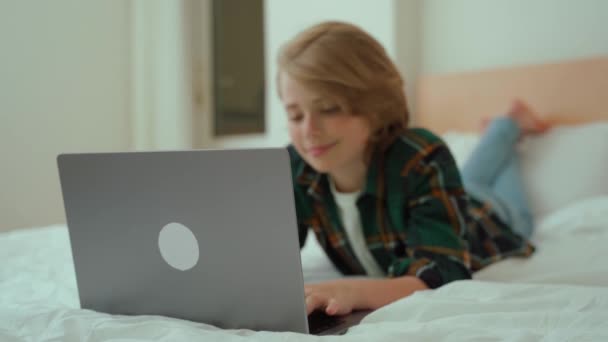 ノートパソコンを使って10代の少年 子供のキーボードのベッドの横に入力します ビデオゲームをプレイする 宿題を勉強している少年 Eラーニング リモートオンライン教育クラスフォアグラウンドに焦点を当てる — ストック動画