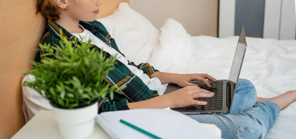 Teenage child boy using laptop. Child typing on the keyboard lying in bed. Play video game. Schoolboy studying doing homework. E-learning, remote educational classes