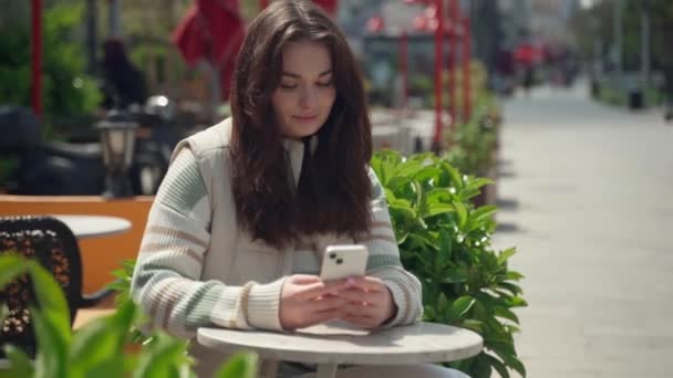 快乐的年轻女子在户外用手机打字 在城市背景下 快乐的少女坐在智能手机旁 社交媒体 暴民应用程序 观看视频 网上购物 浏览互联网 — 图库视频影像