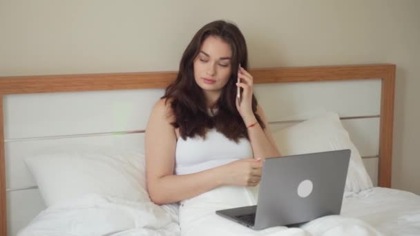 ベッドでノートパソコンを使いながらスマホで話す若い女性 10代の女の子は友人の携帯電話のワーキングコンピュータと話す タイピングを勉強 オンラインショッピング ビデオを見て リール — ストック動画