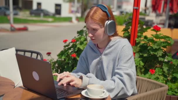在咖啡馆里用笔记本电脑在户外喝咖啡的少女耳机 快乐的年轻女子坐在那里看电视 听着音乐电话 学生们的工作论文 花卉背景的考试研究 — 图库视频影像