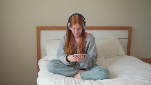 ベッドの上で自宅でスマートフォンを使用して10代の女の子のヘッドフォン 若い女性はガジェットと音楽をオンラインポッドキャストの時計ビデオに耳を傾け 友人とオンラインチャットメッセージインターネット技術 — ストック動画