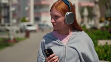 Açık havada akıllı telefon kullanan kızıl saçlı bir kız kulaklığı. Neşeli genç bir kadın yürüyor, müzik dinliyor cep telefonu öğrencisi yaşam tarzı dansından hoşlanıyor.